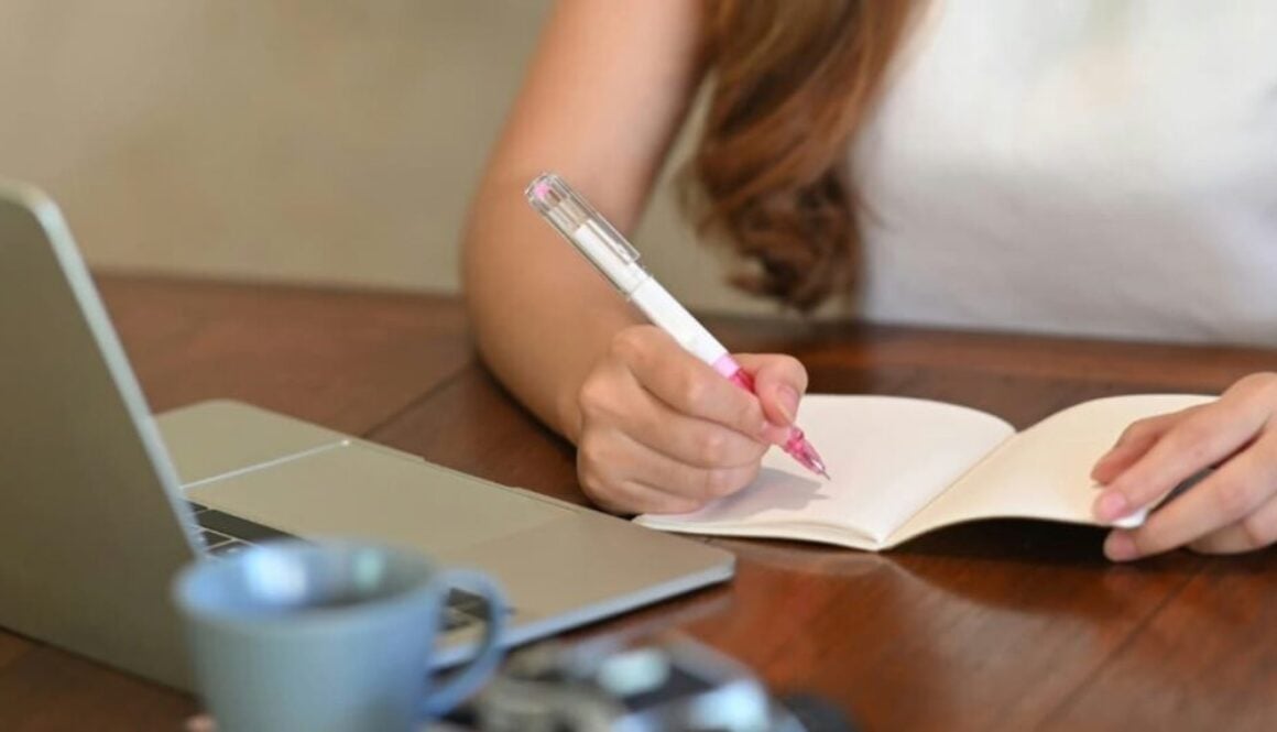 A young woman writes in a notebook while sitting at an open laptop.