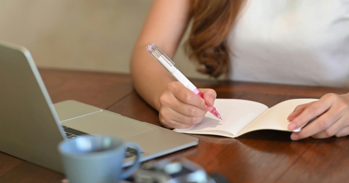 A young woman writes in a notebook while sitting at an open laptop.