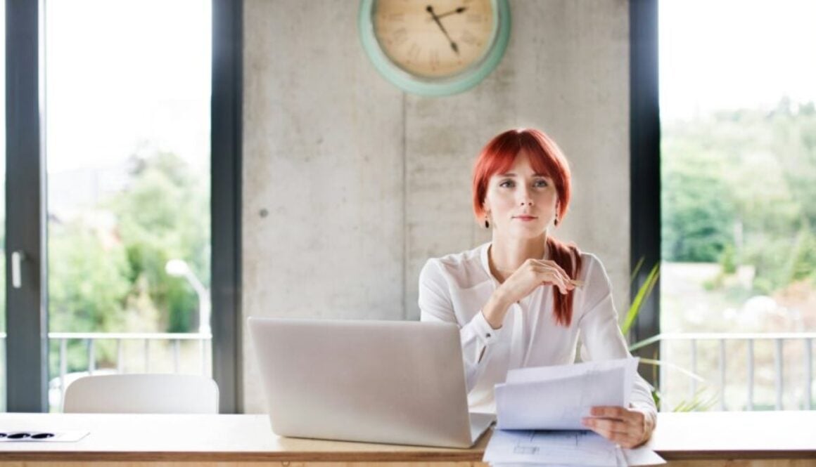 A woman sits thinking at desk in front of papers and a laptop computer.