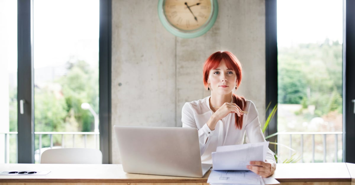 A woman sits thinking at desk in front of papers and a laptop computer.