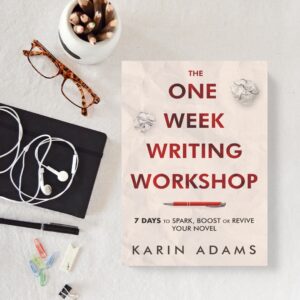 A paperback copy of The One Week Writing Workshop by Karin Adams on a table beside a pencil holder, pair of reading glasses, earbuds and writing supplies.