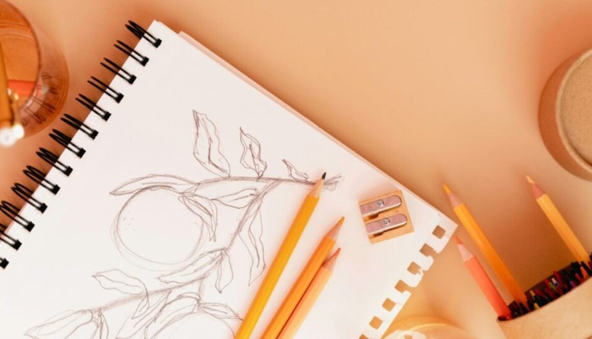 Pencils and a pencil sharpener rest upon an open sketchbook with an image of fruit on a vine.