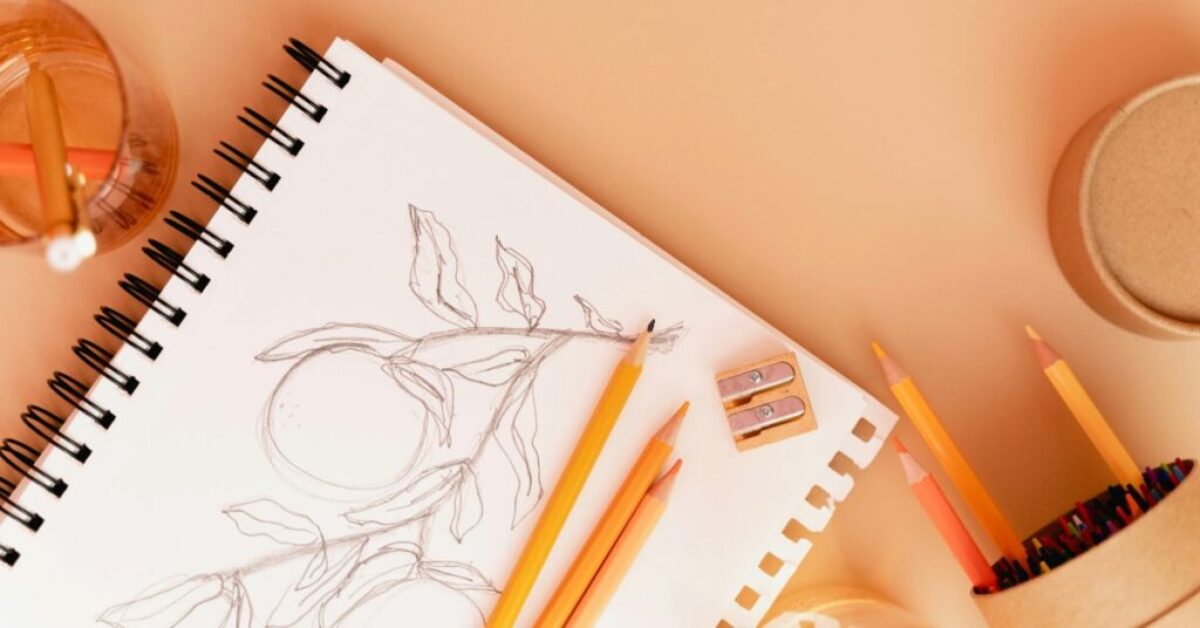 Pencils and a pencil sharpener rest upon an open sketchbook with an image of fruit on a vine.