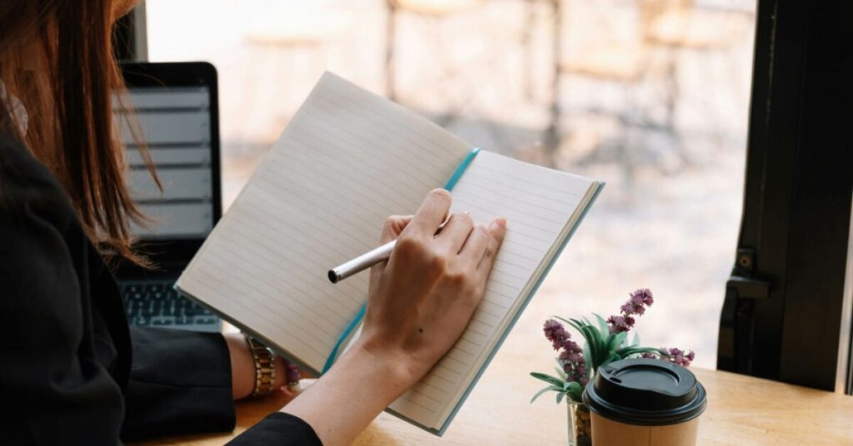 a woman sits at a desk with a cup of coffee and writes in a notebook