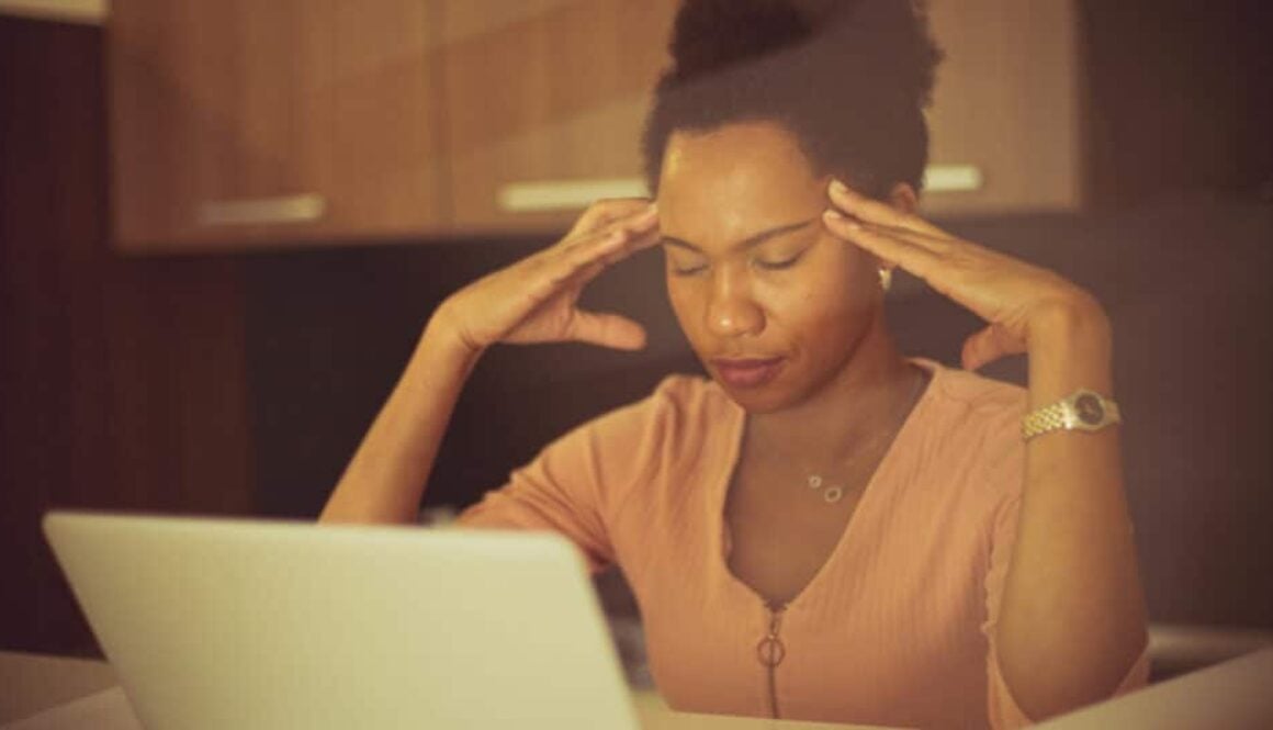 a woman sits at her laptop with eyes closed and fingertips at her temples