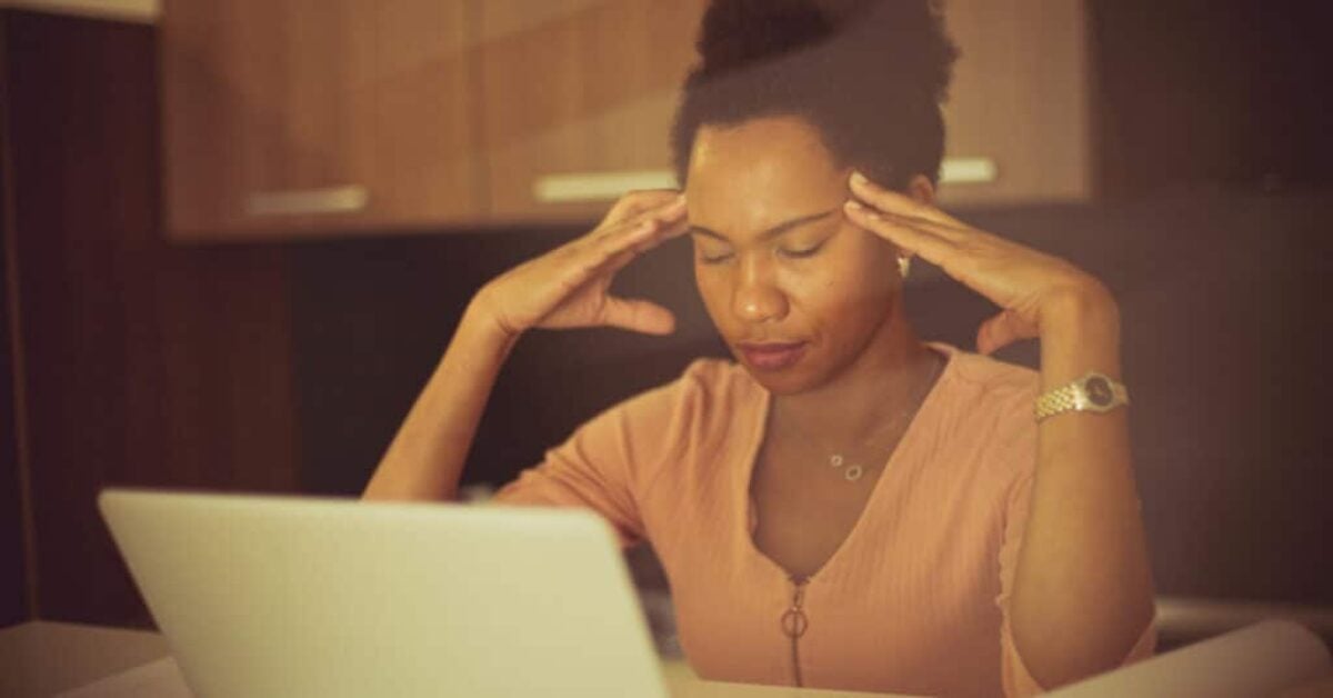 a woman sits at her laptop with eyes closed and fingertips at her temples