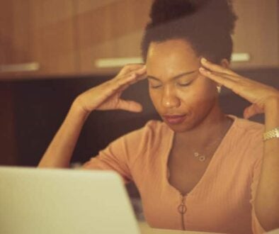 a woman sits at her laptop with eyes closed and fingertips at her temples
