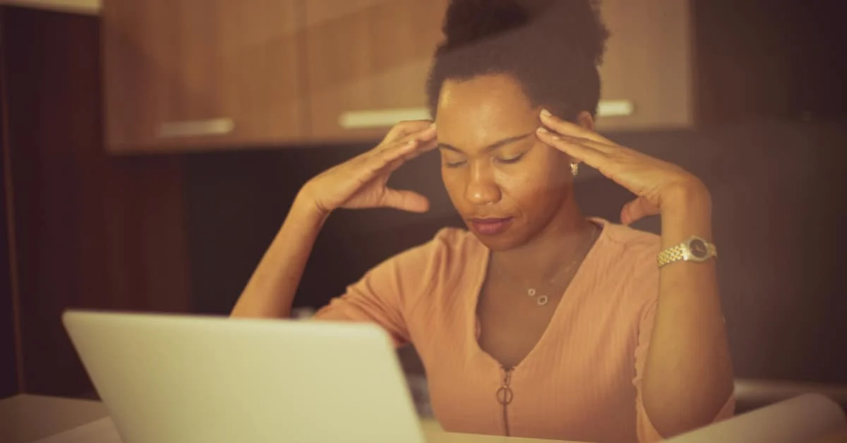 a woman sits at her laptop with eyes closed and fingertips at her temples