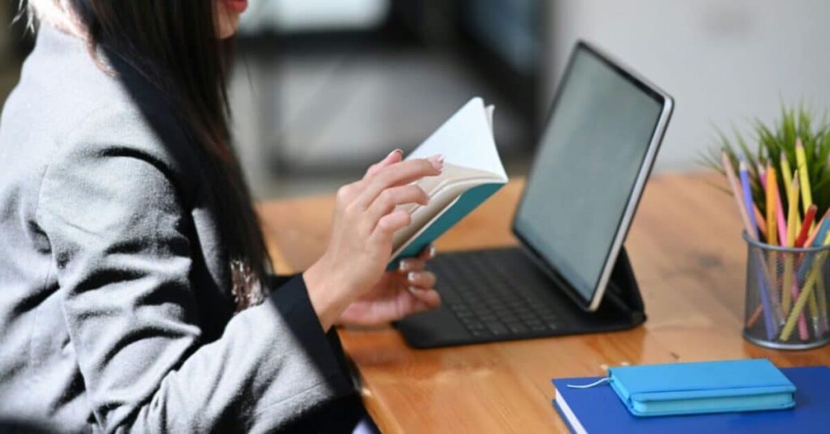 a woman sits at desk in front of an open laptop while flipping through a notebook