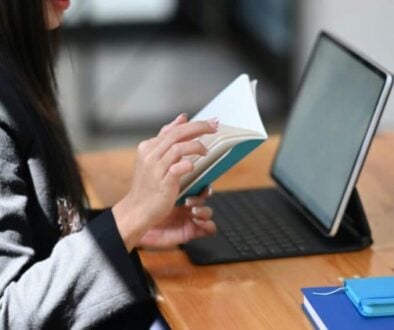 a woman sits at desk in front of an open laptop while flipping through a notebook
