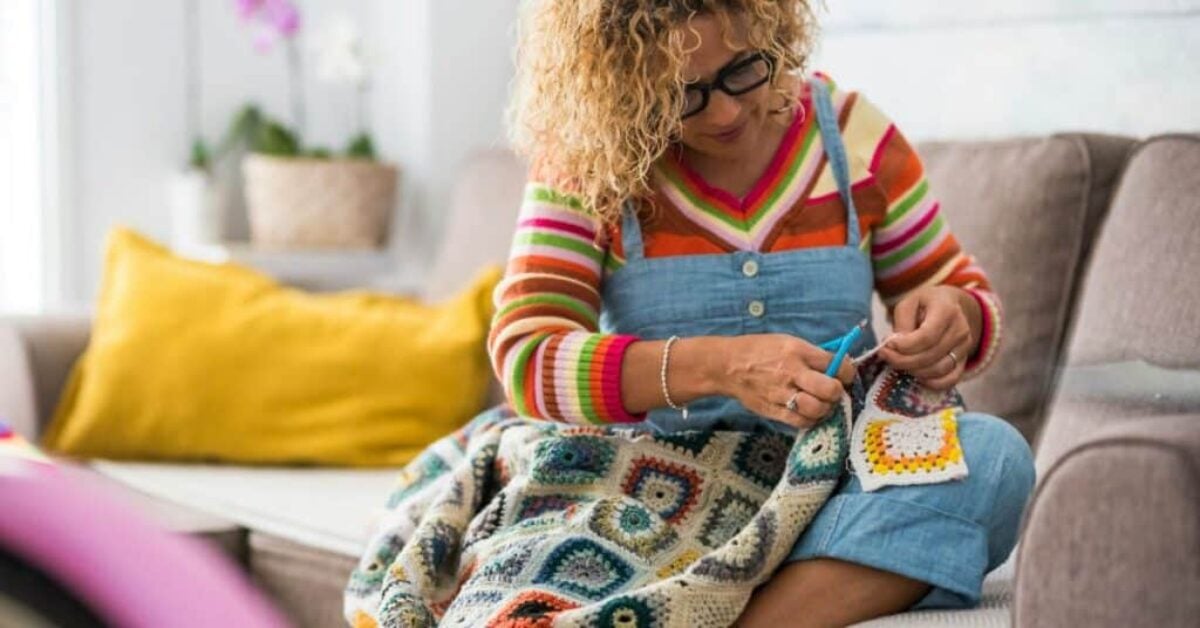 a woman sits on a couch and crochets a blanket