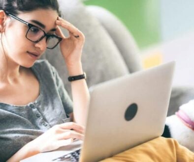 A woman sits on her couch with her laptop and a black and white dog