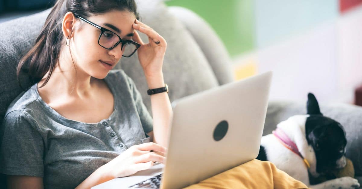 A woman sits on her couch with her laptop and a black and white dog